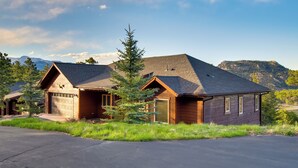 A two-story log cabin with a garage, surrounded by a grassy area and a mountain backdrop. - Lake & Mountain Views: Stunning Estes Park Home! (Estes Park)