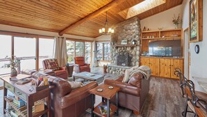 A spacious living area with leather sofas, a stone fireplace, wooden ceiling beams, and a kitchen area with wooden cabinets. - 'Reflections' Cabin on Lake Superior - Near Lutsen (Schroeder)