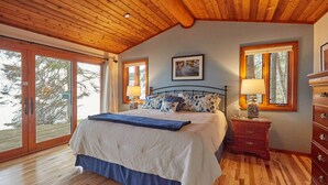 A bedroom with a wooden ceiling, a large bed with floral bedding, two bedside lamps, a dresser, and a large window with a view of trees. - 'Reflections' Cabin on Lake Superior - Near Lutsen (Schroeder)