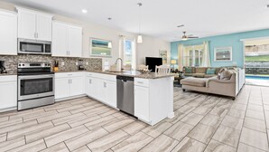 A modern kitchen with white cabinets, a built-in microwave, and a dishwasher. There is a large island with a sink and a flat-screen TV. The room has a light blue wall and a large window with curtains.
