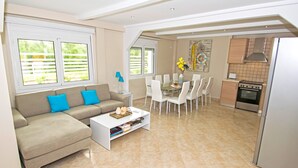 Een moderne woonkamer met een bank, salontafel en een eethoek met stoelen. Er is een keuken met een fornuis en een koelkast.