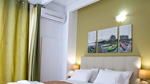 Een hotelkamer met een bed, twee bedlampen, een wandairconditioner en drie schilderijen van boten aan de muur.