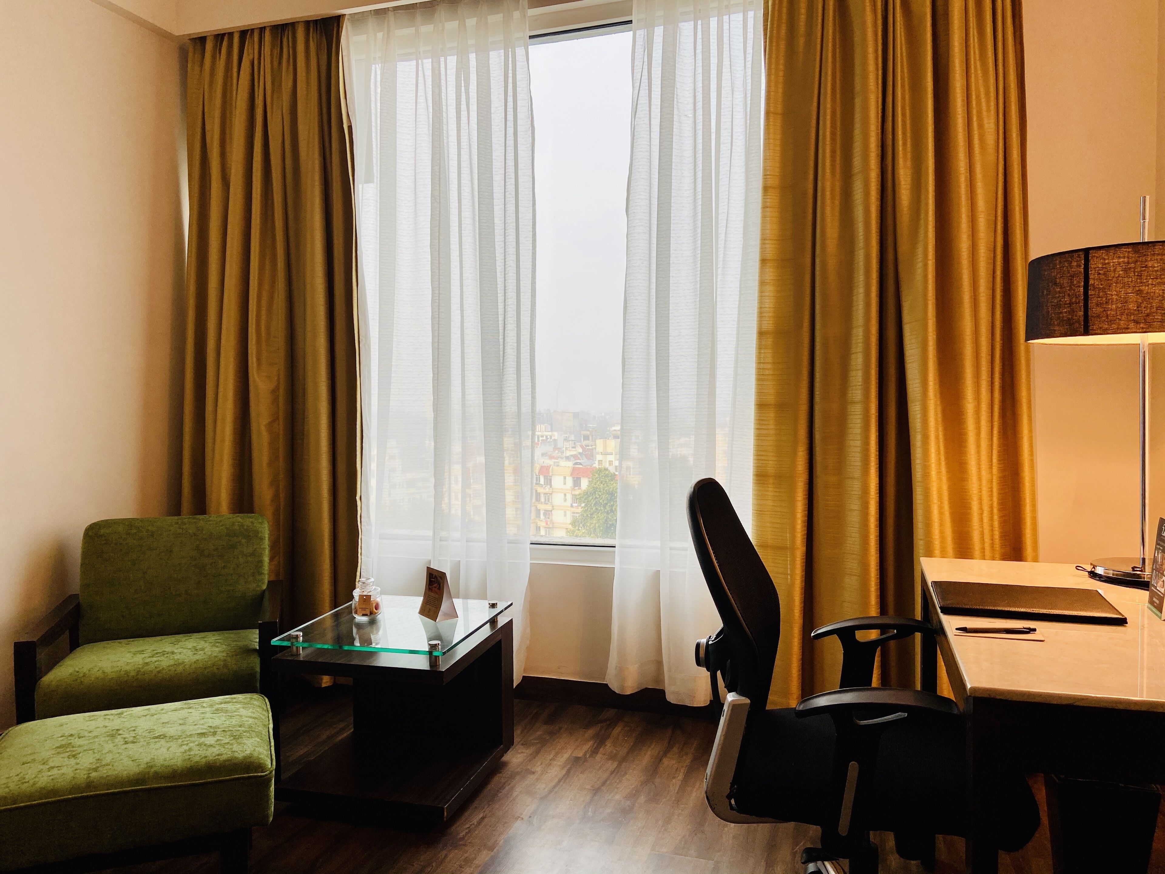 A hotel room with a green armchair, a glass-top table with a picture frame, a black office chair, and a desk with a lamp and a notebook.