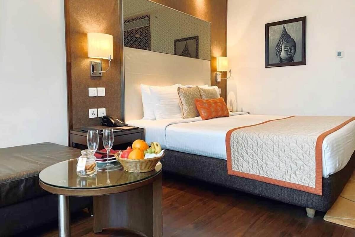 A hotel room with a bed, bedside table with a basket of fruit, a glass table with a bottle and glasses, and a Buddha statue on the wall.