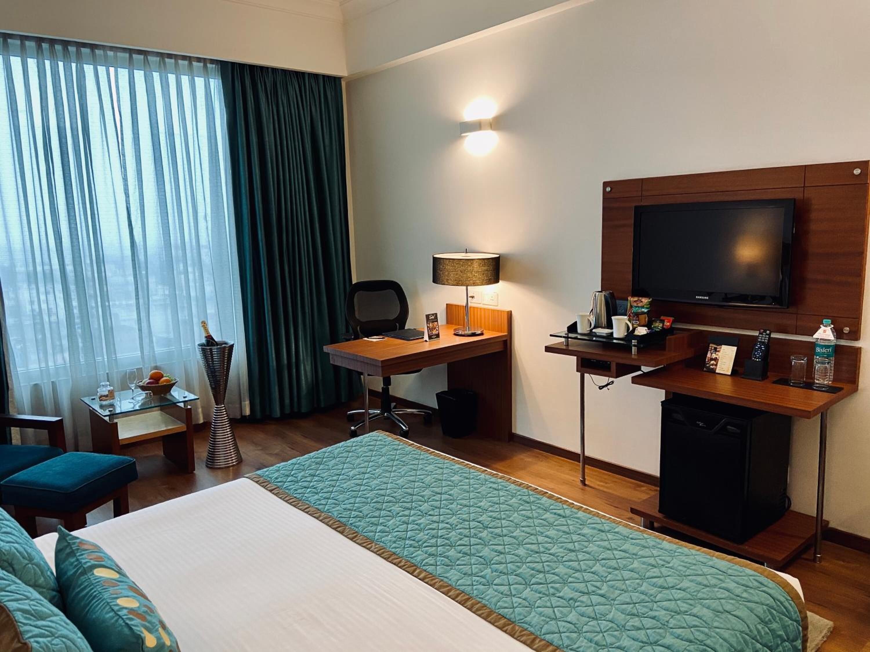 A hotel room with a bed, a desk, a chair, a television, a minibar, and a window with curtains.