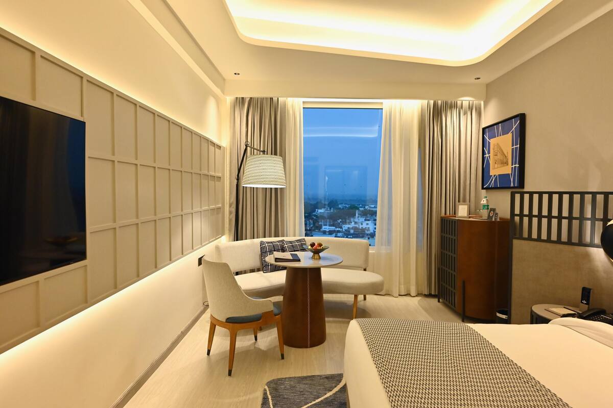A modern hotel room with a sofa, a round table, a TV, a bed, and a view of the city through large windows.