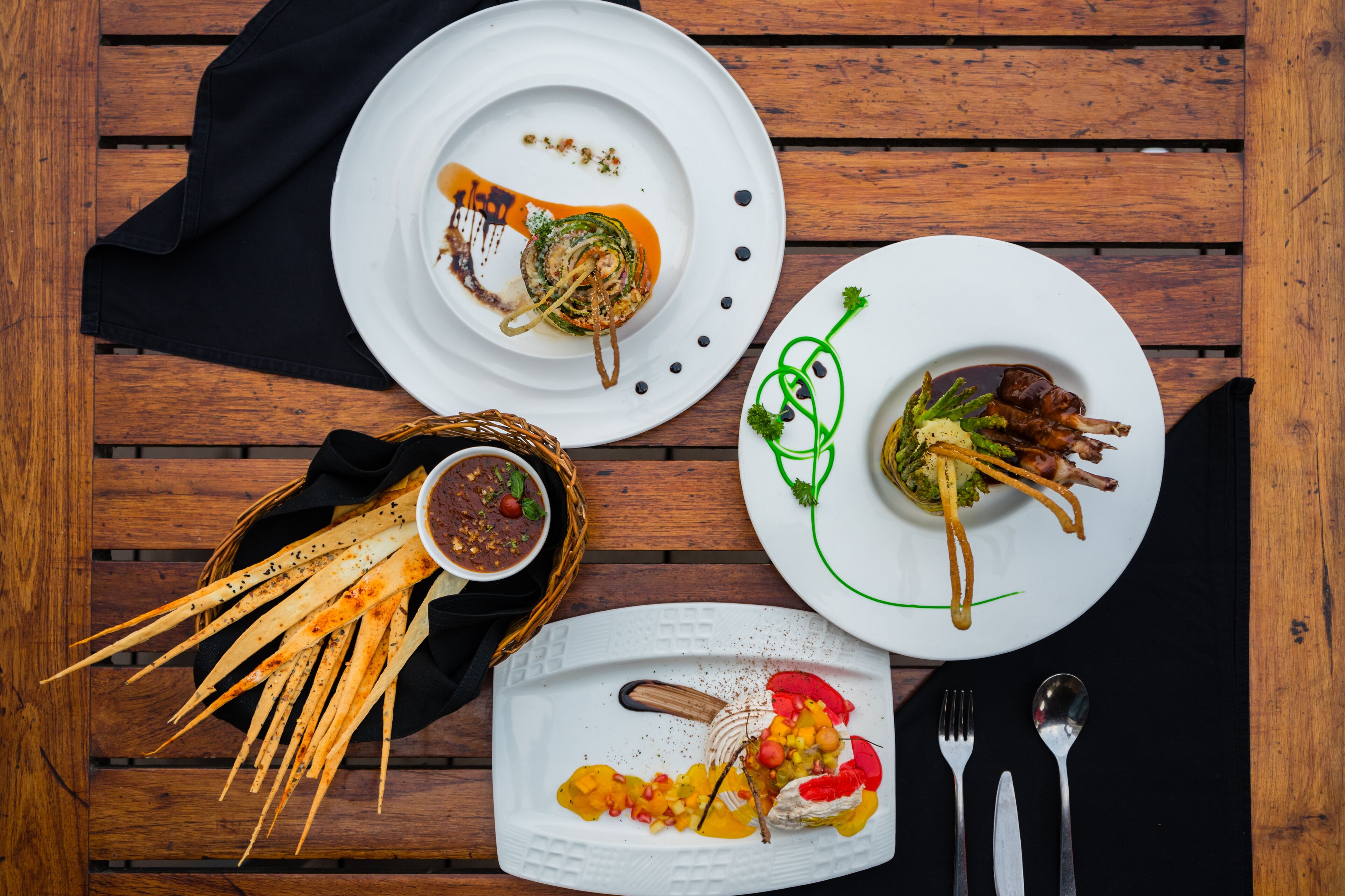 A wooden table with three plates of food, a basket of breadsticks, and cutlery.