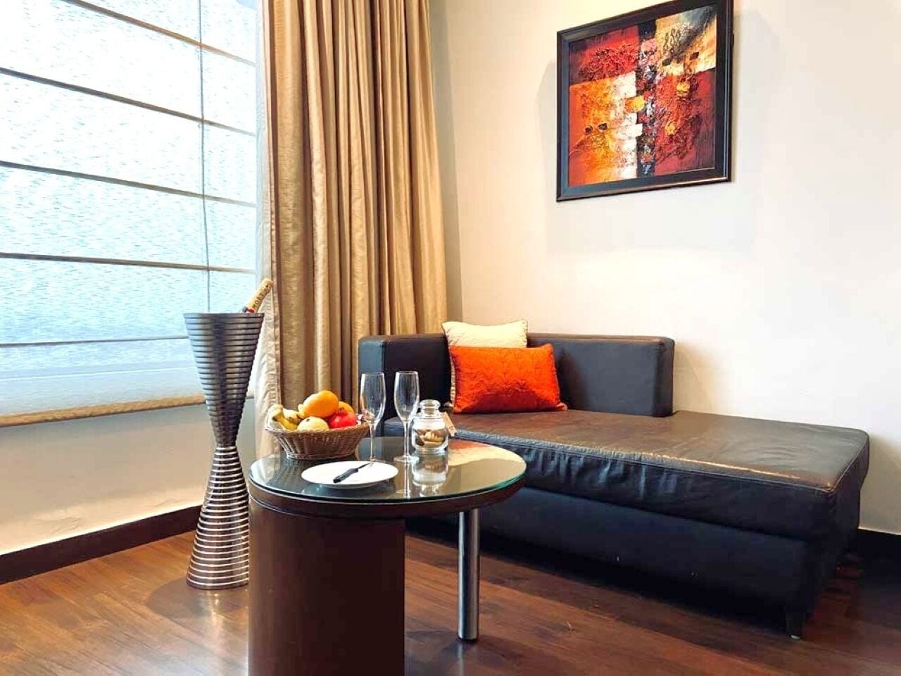 Un salon moderne avec un canapé foncé, une table ronde en verre sur laquelle se trouve un panier de fruits, des verres à vin et un vase.