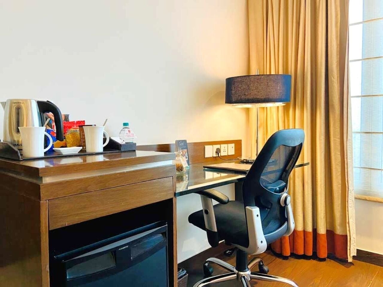 Une chambre d’hôtel avec un bureau en bois, un fauteuil de bureau noir, une lampe, une fenêtre avec des rideaux et une machine à café.