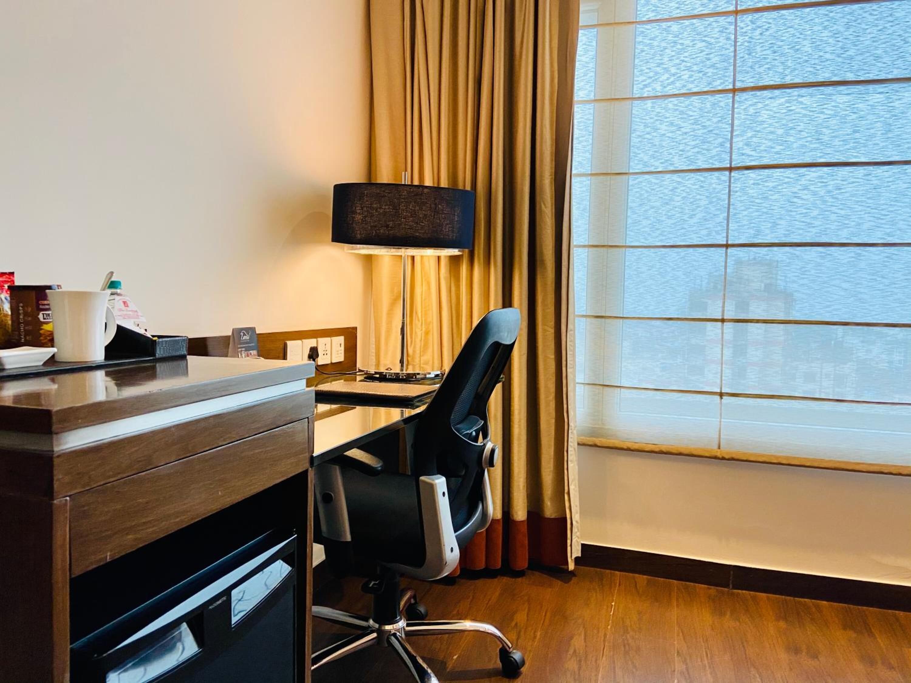 Een hotelkamer met een houten bureau, een zwarte kantoorstoel, een flatscreen televisie, een raam met gordijnen en uitzicht op het water.