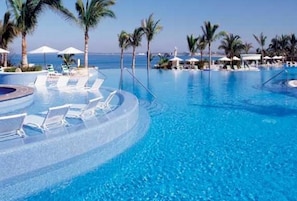 A large, clear blue pool with lounge chairs and palm trees, surrounded by umbrellas and a view of the ocean. - Condo EM (Mazatlán)