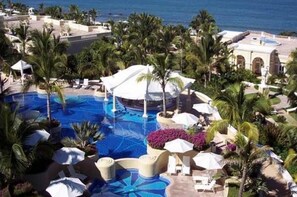A resort pool with palm trees and umbrellas, surrounded by buildings and a view of the ocean. - Condo EM (Mazatlán)