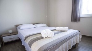 A neatly made bed with white linens and two rolled towels, a bedside table with a cup, and a window with curtains. - Holiday apartment with terrace 200 m from the beach (Primošten)