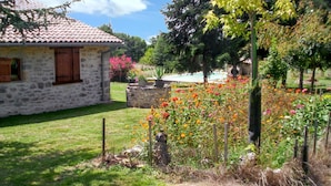 Una casa de piedra con techo de tejas, un patio cubierto de césped y diversas plantas con flores.