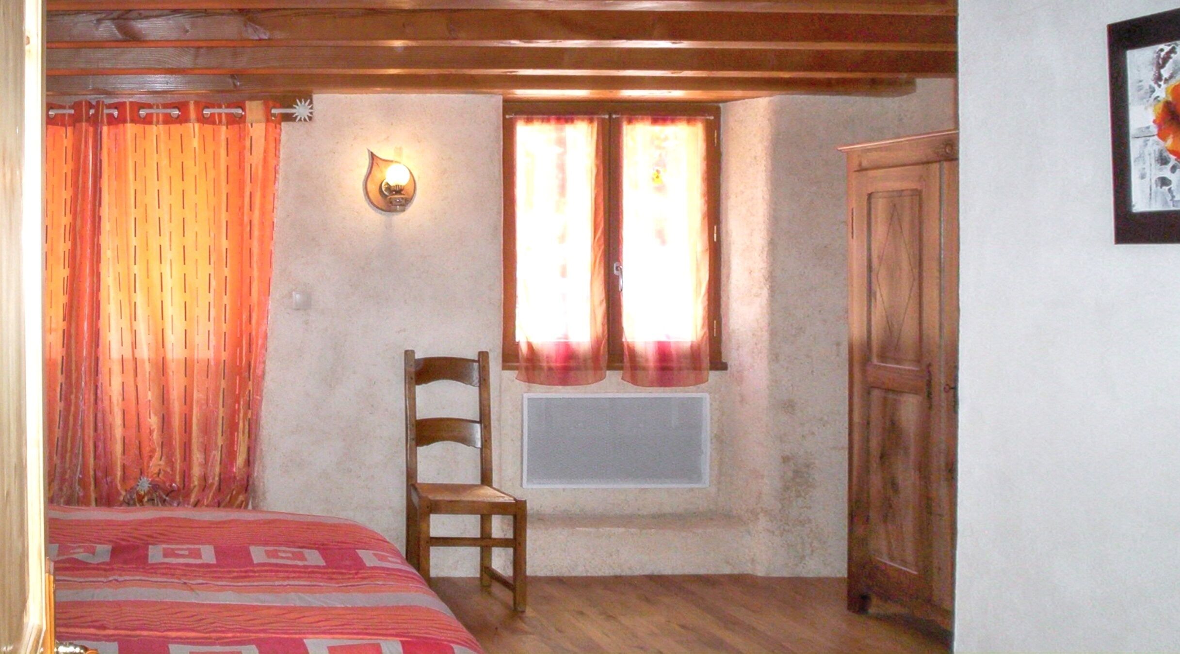 Una habitación con una cama, una silla, un ventanal con cortinas, una lámpara fijada a la pared y un armario de madera.