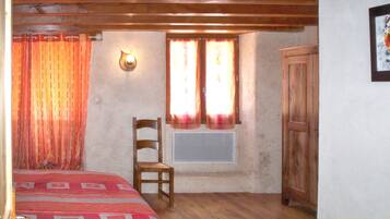 Una habitación con una cama, una silla, un ventanal con cortinas, una lámpara fijada a la pared y un armario de madera.