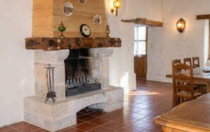 Una chimenea de piedra con repisa de madera, rodeada por un comedor con sillas y mesa de madera.