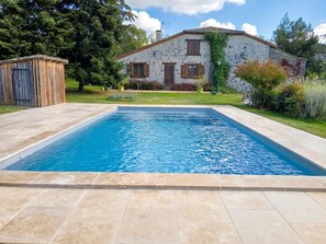 Una piscina rectangular con borde de piedra, un edificio de piedra con contraventanas de madera y una valla de madera al fondo.