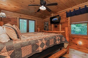 Quarto de cabana de madeira com cama, ventoinha no teto, televisão e janela com vista para árvores.