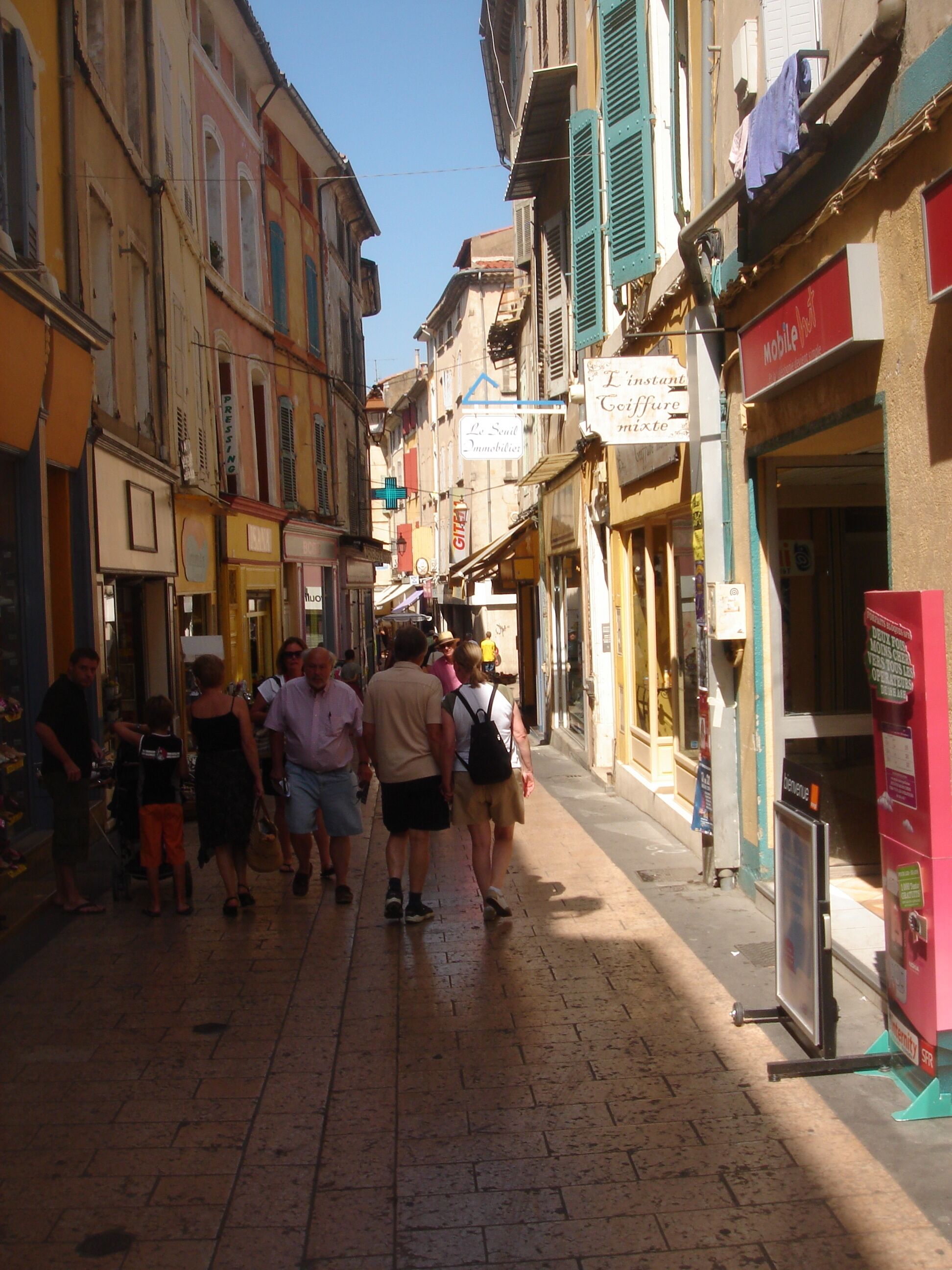 A narrow street lined with colorful buildings, some with signage indicating businesses like a mobile phone store and a press service.