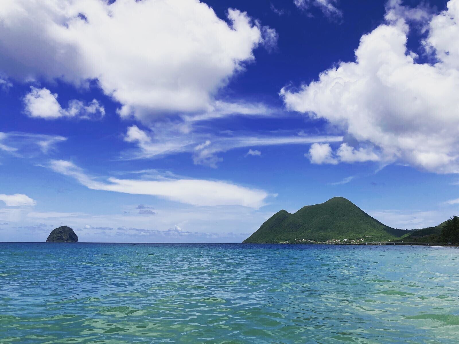 A clear view of a tropical beach with turquoise water, a small island, and a green mountain in the distance.