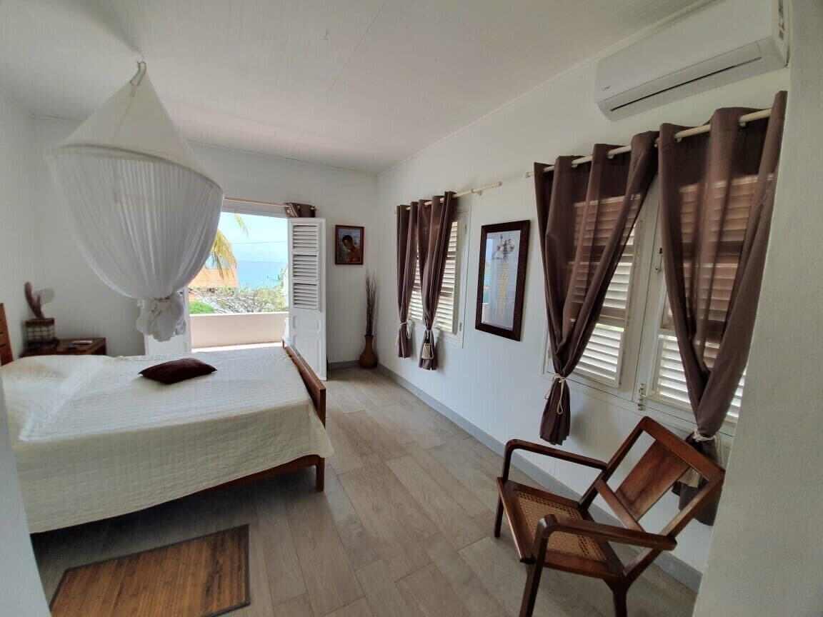 A bedroom with a bed, a chair, a hanging light fixture, a window with curtains, and a door leading to a balcony.