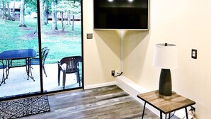A room with a sliding glass door leading to a patio, a flat-screen TV mounted on the wall, a table with chairs, and a floor lamp.