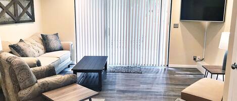 A living room with a sofa, a coffee table, a TV, and a sliding glass door with blinds.