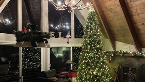 Una stanza accogliente con soffitto alto e travi a vista in legno, e ampie finestre. Al centro, un albero di Natale in bella mostra, circondato da un camino con mensola decorata con addobbi.