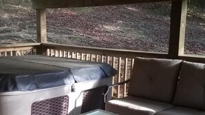Et overdækket udendørs opholdsområde med et bord, en sofa, grill og udsigt til et skovområde.