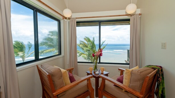 A room with two wooden chairs facing a table, a vase with flowers, and a view of the ocean through large windows.
