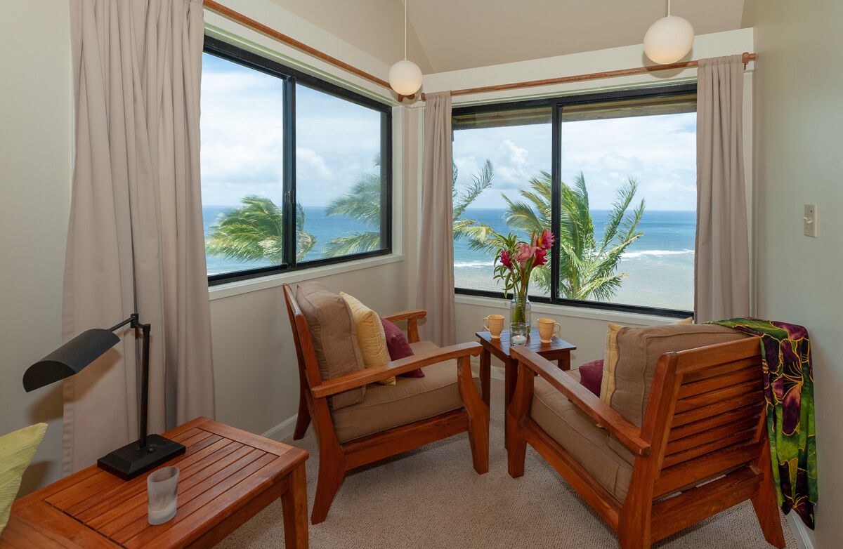 A room with two wooden chairs, a small table, a lamp, and a view of the ocean through large windows.