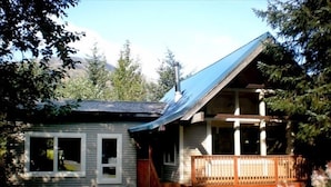 A small cabin with a wooden porch and a steeply pitched roof, surrounded by trees and a mountain backdrop.