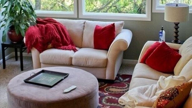 A living room with a beige sofa, a red throw blanket, and a round ottoman. There is a large window with a view of trees.