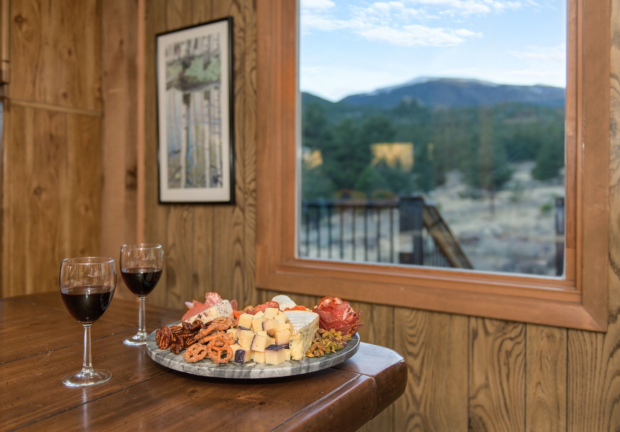 Une table en bois sur laquelle est disposée une assiette de fromages, deux verres de vin rouge, et une vue pittoresque sur les montagnes, visible à travers une fenêtre.