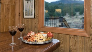 Una mesa de madera con una bandeja de quesos, dos copas de vino tinto y una vista panorámica de las montañas a través de una ventana.