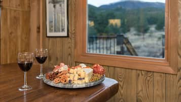 Une table en bois sur laquelle est disposée une assiette de fromages, deux verres de vin rouge, et une vue pittoresque sur les montagnes, visible à travers une fenêtre.