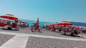 Ein Strand mit Reihen roter Sonnenschirme und weißer Liegestühle, einem klaren blauen Himmel und dem Meer im Hintergrund.