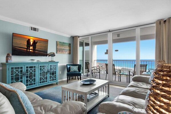 Una sala de estar con un televisor grande, muebles color turquesa y un balcón con vistas al océano.
