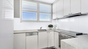 Une cuisine moderne avec des meubles blancs, un évier en acier inoxydable, un lave-vaisselle, une cuisinière et une fenêtre avec des stores.