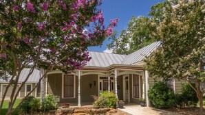 A two-story house with a covered porch, stone pathway, and a well-maintained garden with various plants and a tree with purple flowers. - Herber Haus, Historic Luxury • Walk to Main • Game Room (Fredericksburg)