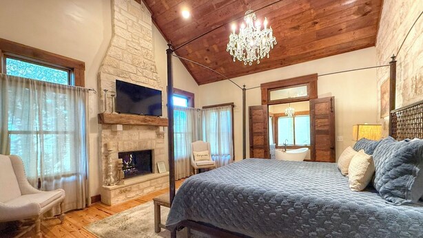 A bedroom with a stone fireplace, a bed with a blue quilt, a chandelier, and wooden beams on the ceiling. - Herber Haus, Historic Luxury • Walk to Main • Game Room (Fredericksburg)