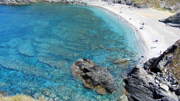 Une plage côtière aux eaux turquoise et limpides, bordée de rochers et d’une étendue de sable.