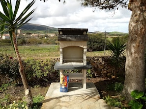 Un horno de piedra al aire libre con un saco de carbón cerca, rodeado de follaje y un muro de piedra.
