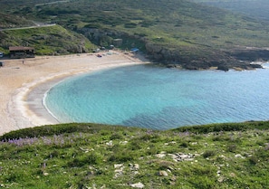 Un paisaje costero con una playa arenosa, aguas azules cristalinas y una colina cubierta de hierba en primer plano.