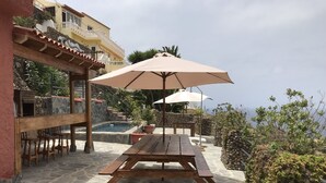 Un patio in pietra con tavolo e panche in legno, una piscina esterna e vista su un edificio con un balcone.