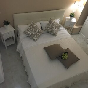 Un letto rifatto con lenzuola bianche e cuscini decorativi, un comodino con una pianta e un abat-jour.