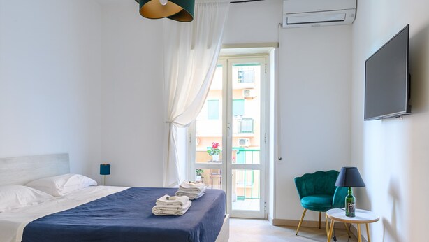 A bedroom with a bed, a chair, a table with a lamp, and a TV on the wall. - Barsanti Apartment (Roma)