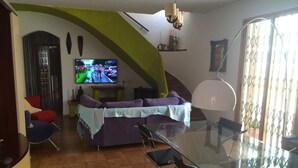 Ein Wohnzimmer mit einer Couch, Sesseln, einem Glastisch und einem Fernseher, der ein Sportereignis zeigt.