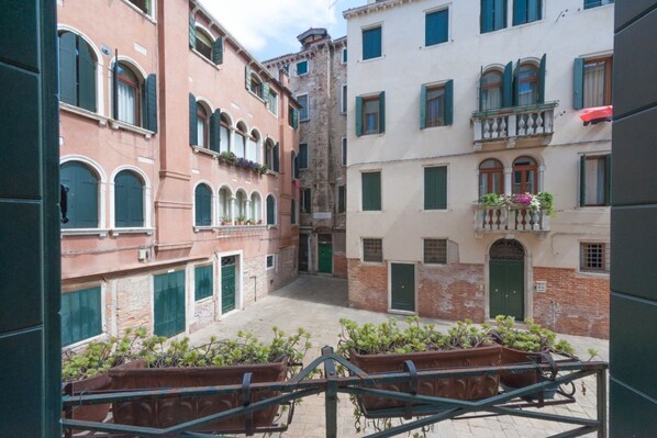 A narrow street with buildings on either side, featuring balconies with plants and a central open space.